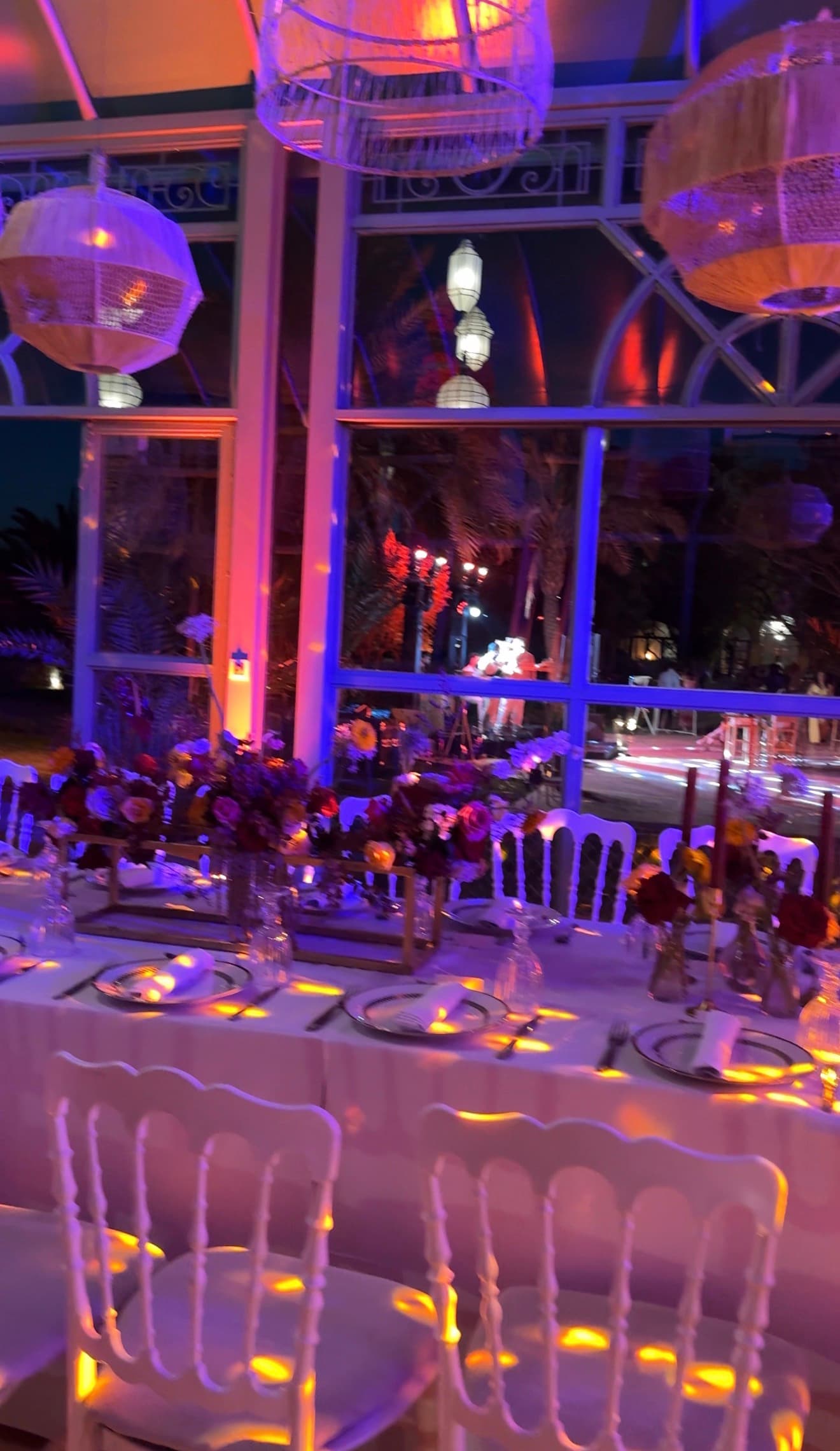 Wedding reception under a glass pavilion, with rattan pendants, flowers and white chairs