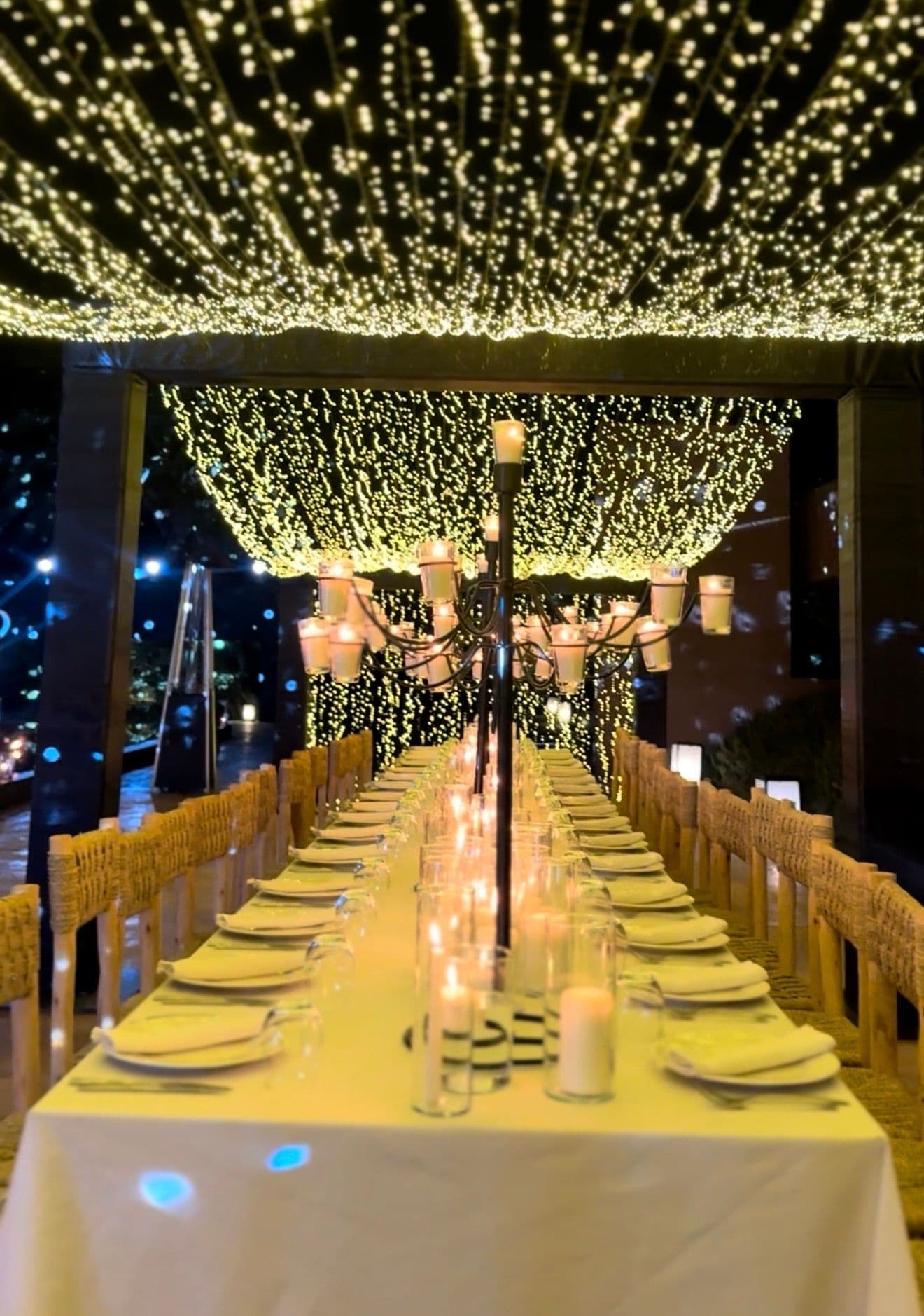 Longue table de dîner sous un rideau de lumières, reflétée dans un bassin aux bougies flottantes